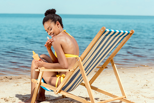  african american woman applying sunscreen lotion on skin while sitting on deck chair on sandy beach